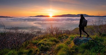 Image of a male stood on a rock at the top of a mountain overlooking a landscape covered in cloud