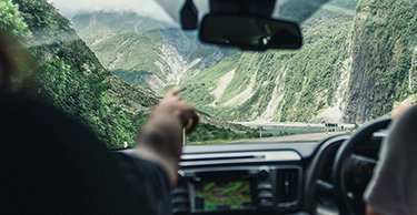 Image of a hand pointing forward from the front seat of a car as two people drive down a steep road in the South Island