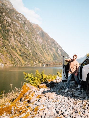 Two men unpacking a car by a mountain lake during a scenic road trip