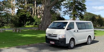 Side image of a Toyota Hiace 10-seater minibus parked in a New Zealand park next to the grass under a tree